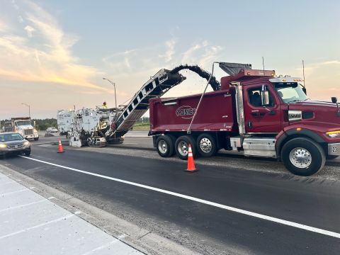 A milling machine discharging asphalt millings into a dump truck on a highway at dawn